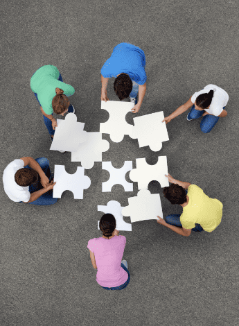 From above we see 5 people with large white puzzle pieces, fitting them together.